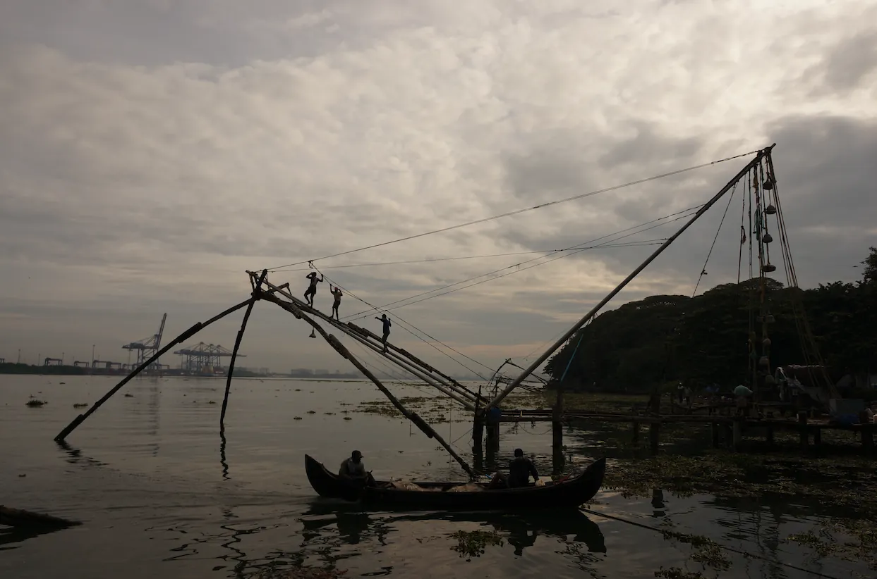 Silhouetted fishermen balancing on the frame of a large cantilevered fishing net at dusk.