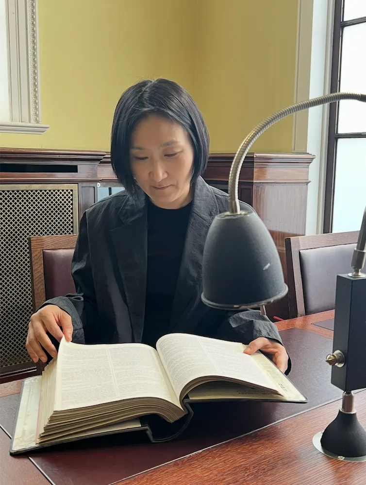 Loonie Park reading a large open book at a wooden library desk under a gooseneck lamp.